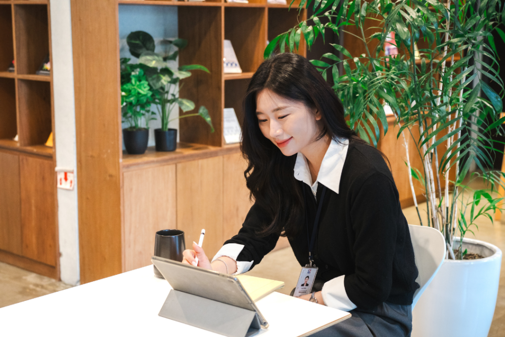 หญิงสาวชาวเอเชียกำลังใช้ปากกา Stylus เขียนหรือทำงานบนแท็บเล็ตในพื้นที่ทำงานสีอบอุ่น