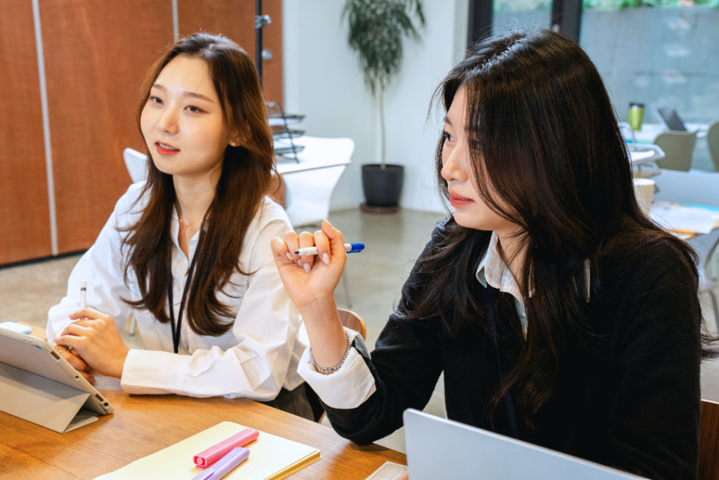 หญิงสาวสองคนกำลังหารือกันอย่างจริงจังในที่ประชุม โดยมีแล็ปท็อปและแท็บเล็ตอยู่บนโต๊ะ