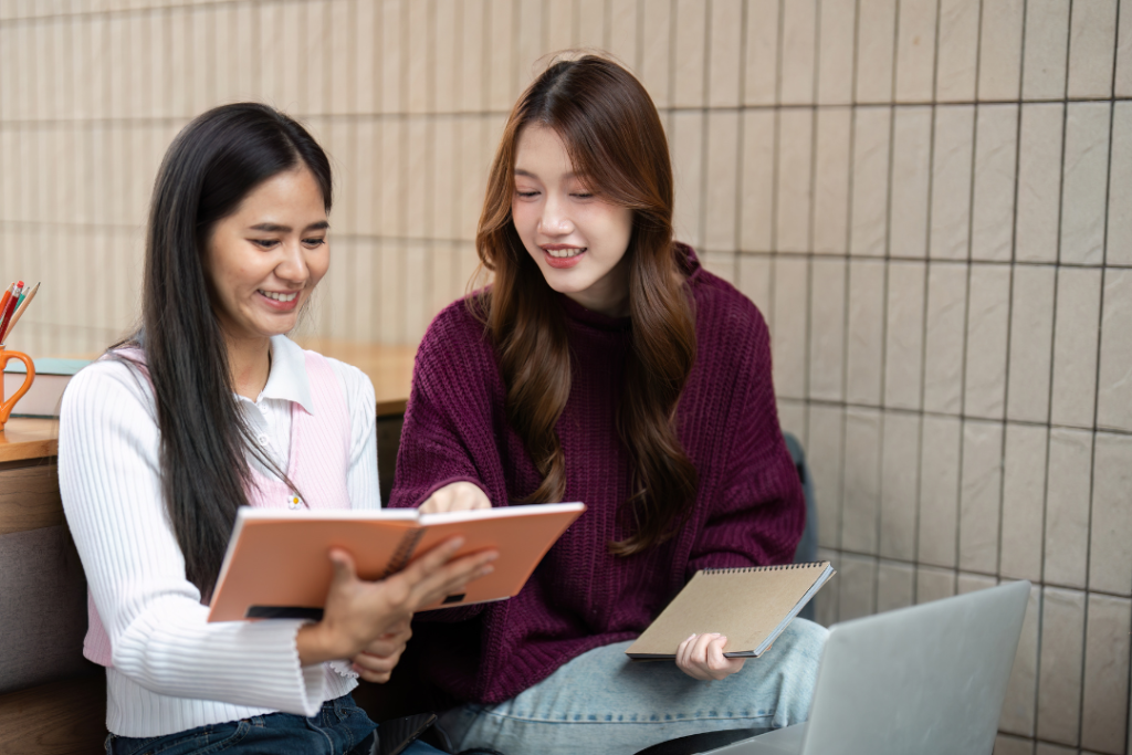 หญิงสาวสองคนกำลังทำงานร่วมกันและชี้ไปที่สมุดโน้ตบนโต๊ะเพื่อแลกเปลี่ยนความคิดเห็น