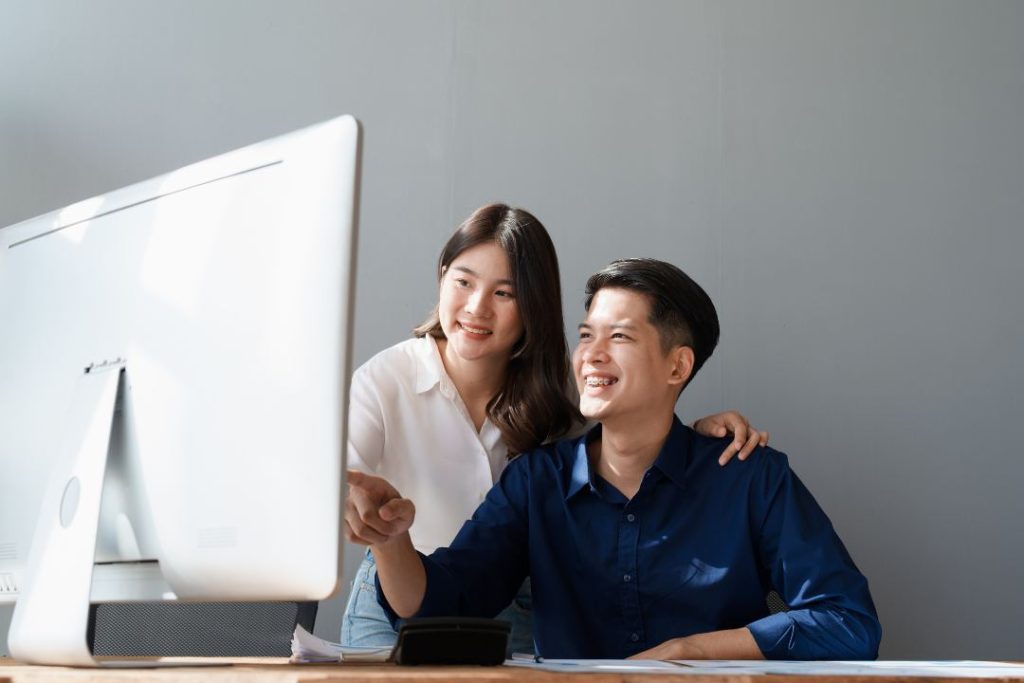A professional Asian man and woman in a modern, bright, windowed office are smiling and looking at a laptop together. They are wearing ID lanyards and appear to be discussing business data or brand performance metrics. There are charts on the desk. This symbolizes Top Reach Marketing’s collaborative and data-driven approach to measuring brand health for clients.