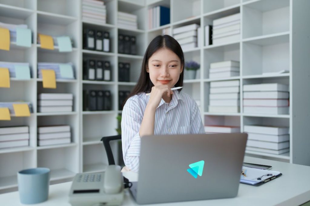 A professional young Asian female marketer sitting at a tidy white desk in a modern office library, looking thoughtfully at a laptop screen with the Top Reach Marketing logo. She is holding a stylus to her chin, representing the strategic thinking and analysis required for Demand Generation and Target Account List planning.