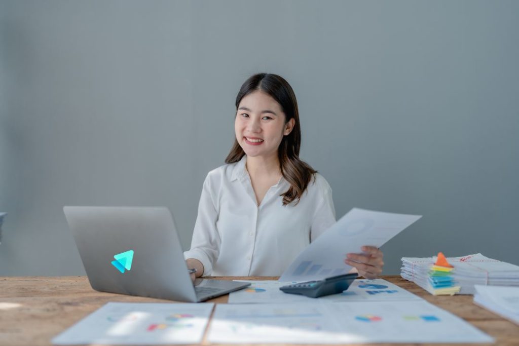An Asian woman in a professional white shirt is smiling and pointing at a large desktop computer screen, likely a chart or map. A man in a blue shirt stands next to her, smiling broadly and looking at the screen. Sunlight from a window illuminates them. They are in a modern, professional office environment. Her gesture symbolizes identifying high-opportunity locations based on market data for Geographic Expansion.
