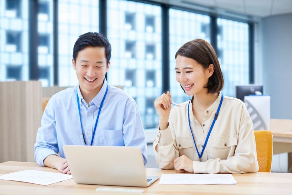 Two professional marketing colleagues smiling and reviewing customer data on a laptop in a modern office, symbolizing marketing and support team collaboration to optimize the Cost of Servicing (COS).