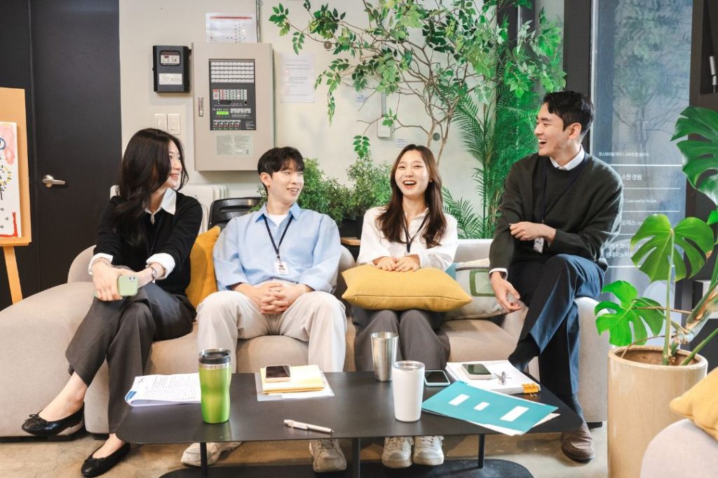 A group of four diverse young Asian business professionals in casual office wear sitting happily on a sofa in a modern, plant-filled collaborative workspace. They are smiling and laughing while having a discussion, with laptops, tablets, and drinks on the table. This represents successful strategic partnership, collaboration, and shared business growth driven by data.